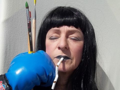 Frau mit schwarzer Perücke hat eine lange Zigarette im Mund und bekommt einen Boxhandschuh mit Pinseln ins Gesicht gehalten
