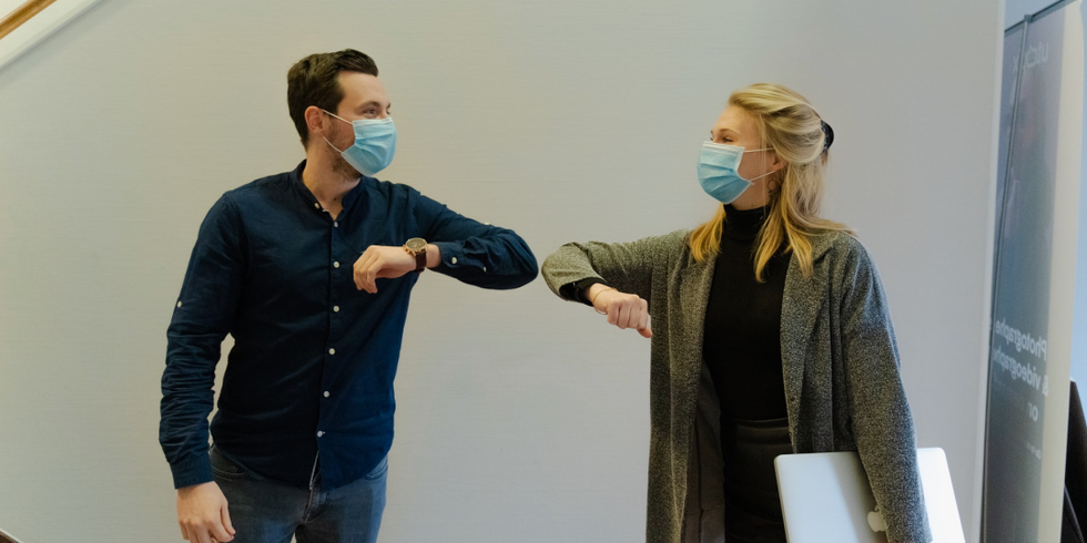 Eine Frau und ein Mann tragen eine medizinische Maske und begrüßen sich mit einem Ellbogencheck.