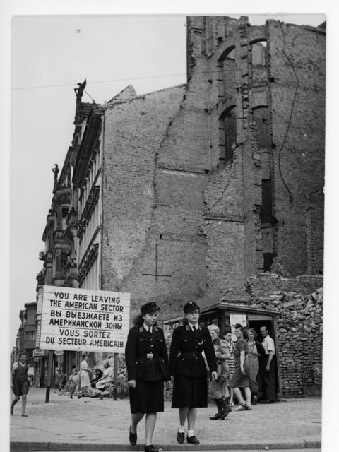 Enlarge photo: Two young women in uniforms cross a road in front of a destroyed building.