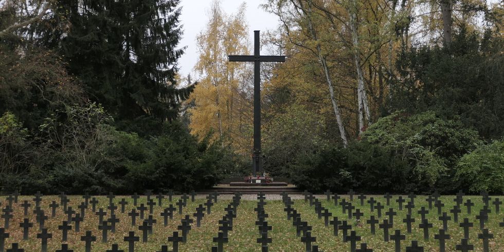 Auf einem Friedhof stehen viele kleine Grabkreuze auf einem Platz. Im Hintergrund steht ein großes Kreuz in der Mitte.