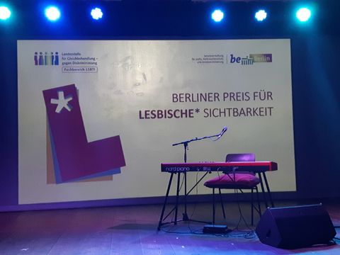 Bildvergrößerung: Bühne mit Mikro und Keeboard, im Hintergrund steht: Berliner Preis für lesbische* Sichtbarkeit