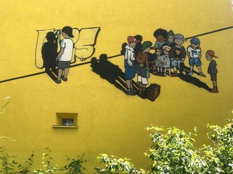 Bildvergrößerung: An einer gelben Hauswand ist ein Gemälde von mehreren Kindern. Ein Kind mal ein Graffiti an eine Wand.