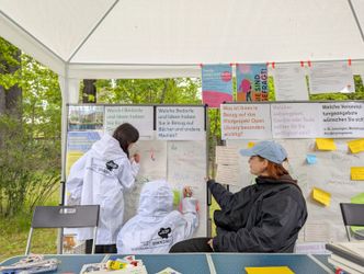 Zwei Kinder in weißen Regenponchos mit dem Logo der Kontaktstelle Beteiligung (Raum für Beteiligung Spandau) schreiben an Pinnwänden mit Fragen zur neuen Stadtbibliothek, eine Mitarbeiterin sitzt daneben und assistiert, im Hintergrund hängen bunte Plakate.