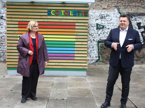 Bildvergrößerung: Eine Frau und ein Mann stehen vor einer Holzwand, die in den Farben des Regenbogens bemalt ist.