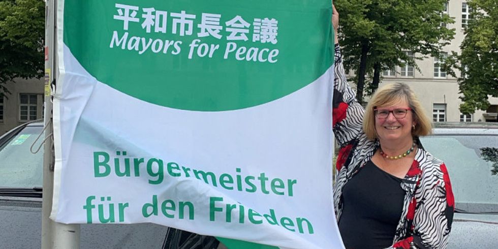 Eine Frau hält einen Flagge mit der Aufschrift "Mayors for Peace, Bürgermeister für den Frieden" an einem Mast hoch.