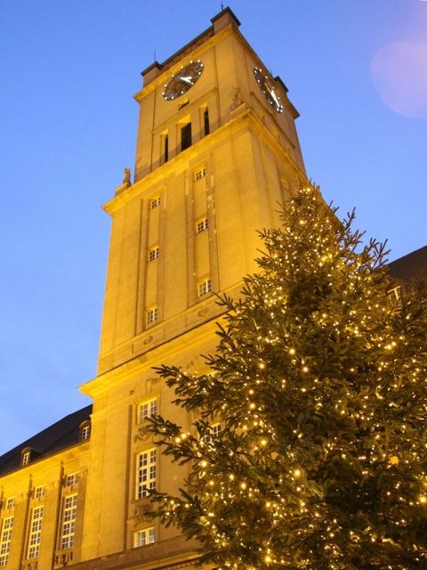 Beleuchteter Weihnachtsbaum vor einem Turm bei Dämmerung.