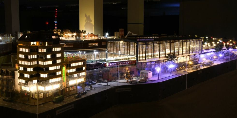Eine Modelleisenbahn in einem abgedunkeltem Raum mit vielen Lichtern an kleinen Gebäuden und Straßenlaternen. Im Vordergrund ist eine Bahnhofshalle und im Hintergrund ist der Fernsehturm.