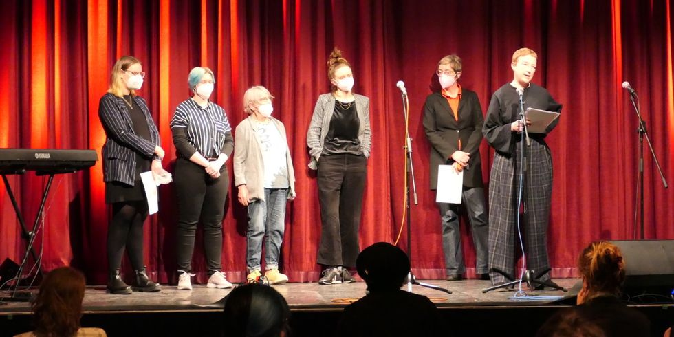 Auf einer Bühne vor einem roten Vorhang stehen 6 Frauen. Eine Frau steht ein Stück weiter vorne und spricht in ein Mikrofon.