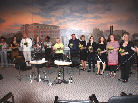 Bildvergrößerung: 12 feierlich gekleidete Erwachsene stehen nebeneinander mit einem Blumenstrauß in der Hand. Rechts steht eine Frau und spricht ins Mikrofon