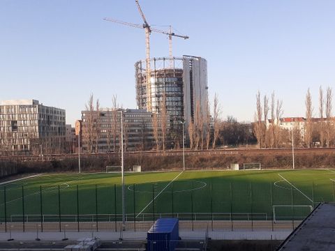 Ein grüner Fußballplatz vor einem Gleis. Im Hintergrund ist das Schöneberger Gasometer, ein bebauter Stahlgerüst-Zylinder, mit zwei Baukränen.