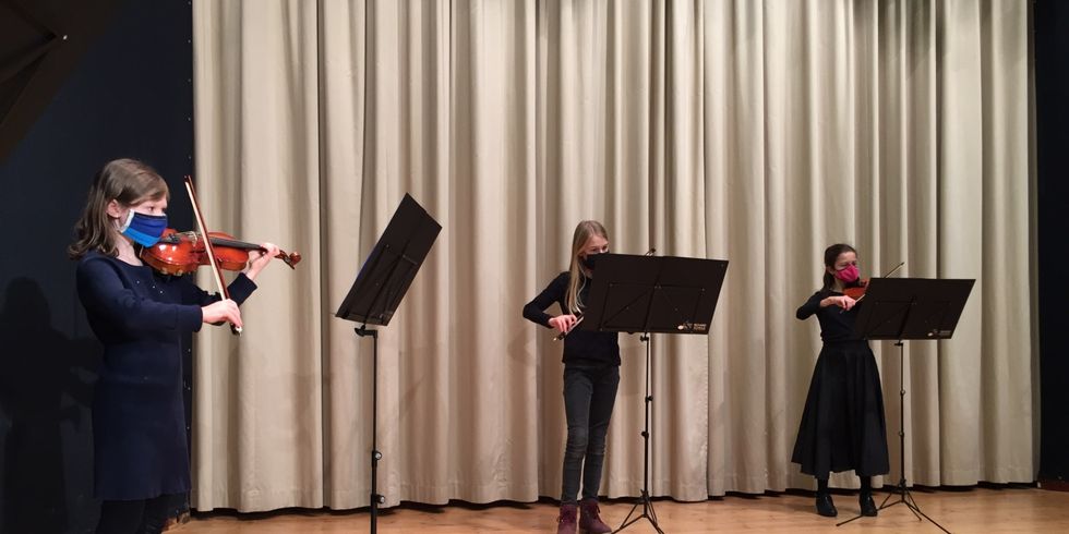 drei Kinder stehen in großem Abstand auf einer Bühne und spielen Geige 