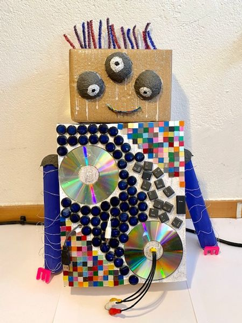 Bildvergrößerung: Ein kleiner selbst gebastelter Roboter mit einem Kopf aus Passe und drei Augen aus Styropor sitzt gegen eine Wand gelehnt und lächelt.
