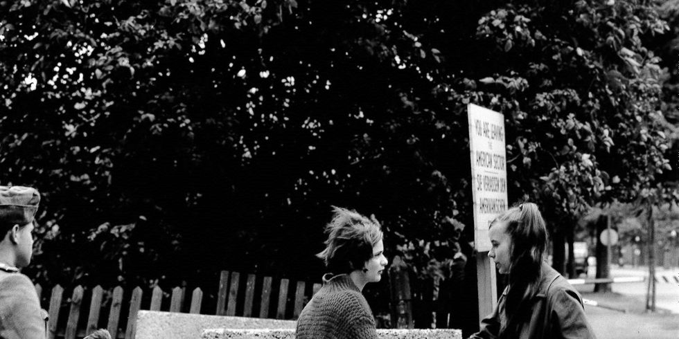 Zwei Mädchen an der noch nicht fertiggestellten Berliner Mauer in Treptow in der Harzer Straße. Sie sprechen miteinander und fassen sich an. Etwas weggedreht steht ein Soldat. Er hat keine Pistole und kein Gewehr in der Hand, sondern eine Blume.