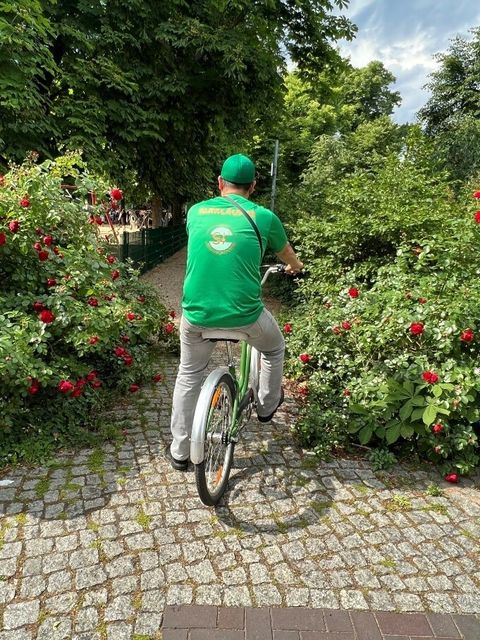 Ein Mann in einem grünen T-Shirt und einer grünen Kappe sitzt auf einem grünen Fahrrad zwischen blühenden Sträuchern.