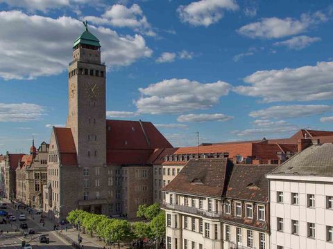 Im linken Bildteil das Rathaus Neukölln. Heller Sandsteinbau mit rotem Dach und einem Rathausturm mit einem grünen Dach. Davor eine Häuserzeile