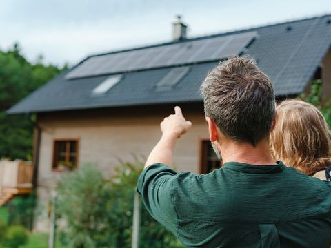 Mann mit Kind auf dem Arm vor einem Einfamilienhaus mit Solaranlage auf dem Dach