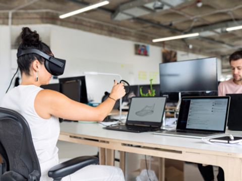 Frau mit VR-Brille vor Laptop, ihr gegenüber sitzt ein Mann