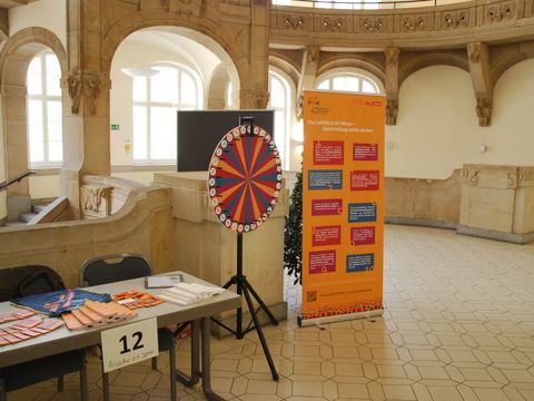 Bildvergrößerung: Ein Tisch mit Infomaterial der Kampagne, das Glücksrad und ein Roll-Up in der Rathausgalerie Charlottenburg-Wilmersdorf