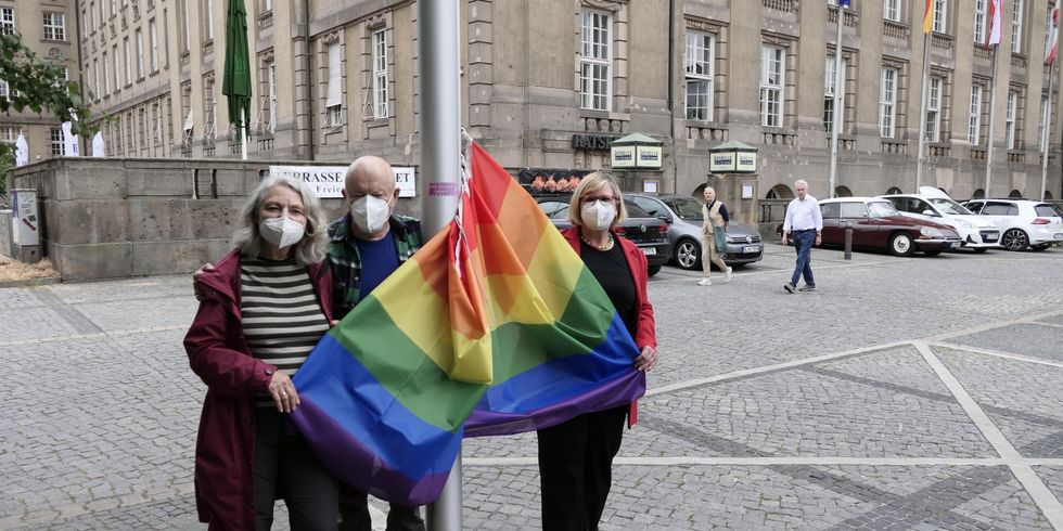 Drei Personen mit Masken halten eine Regenbogenflagge, die an einem Fahnenmast hängt.