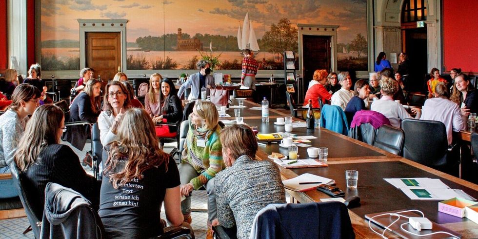 In einem großen Saal mit Wandmalerei sind Frauen an Tischen und in Stuhlkreisen und unterhalten sich miteinander.
