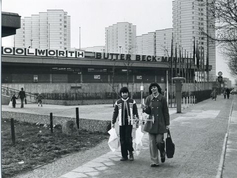Historische Ansicht der Gropiusstadt. im Vordergrund zwei Frauen mit Einkaufstaschen. Im Hintergrund ein Gebäude mit dem Schriftzug Woolworth. Dahinter Hochhäuser der Gropiusstadt