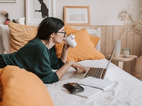 Eine junge Frau liegt auf dem Bett mit einer Tasse und arbeitet am Laptop