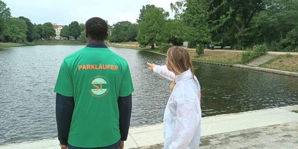 eine Frau und ein Mann stehen an einem See, Der Mann trägt ein T-Shirt mit der Aufschrift "Parkläufer"
