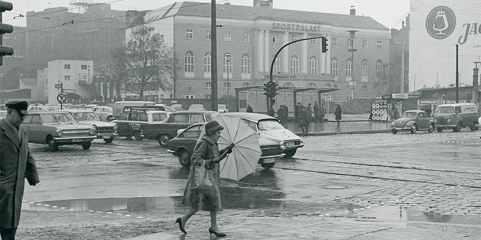 Schwarzweiß-Fotografie von einer Kreuzung an einem regnerischen Tag. Im Vordergrund läuft eine Frau mit einem Regenschirm auf dem Gehweg und im Hintergrund steht an einem großen Gebäude der Schriftzug "Sportpalast".