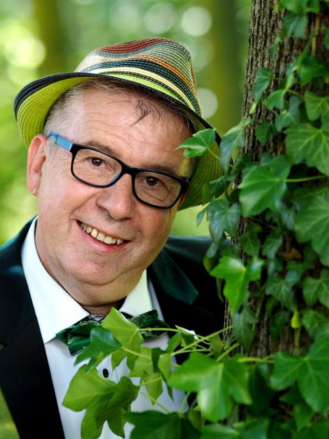 Ein lächelnder Mann mit Hut und Brille schaut hinter einem mit Efeu-bewachsendem Baum hervor.