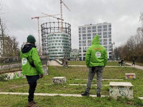 Bildvergrößerung: Auf einer Grünfläche stehen im Vordergrund zwei Menschen mit grünen Jacken. Sie schauen in die Ferne. Dort steht ein Stahlgerüst, das Schöneberger Wahrzeichen, der Gasometer.