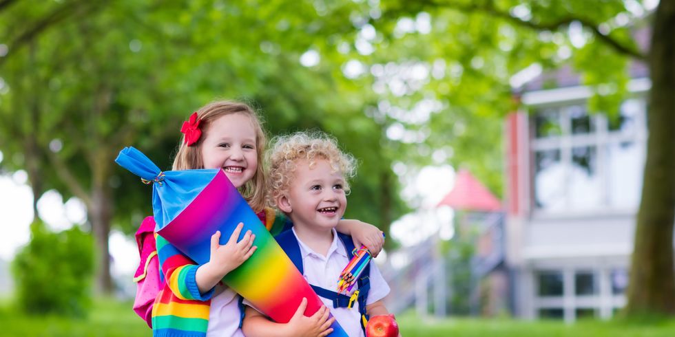 Zwei lachende Kinder mit einer regenbogenfarbenen Schultüte im Arm auf einer Wiese