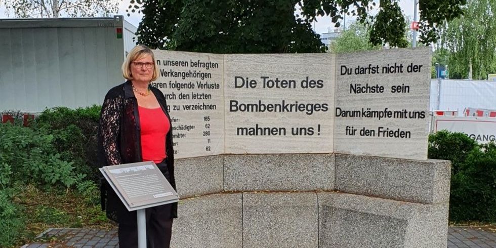 Eine Frau steht neben einem Denkmal aus Stein mit der Inschrift "Die Toten des Bombenkrieges mahnen uns!". Vor ihr ist eine kleine Informationstafel aufgestellt.