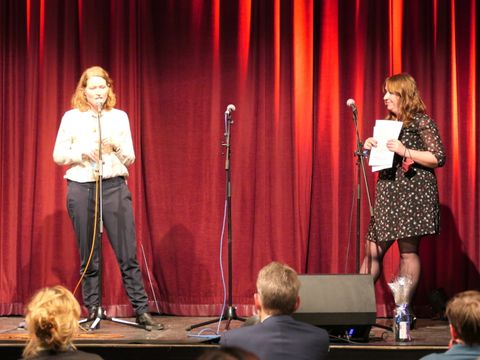 Bildvergrößerung: Zwei Frauen stehen auf einer Bühne vor einem roten Vorhang. Beide stehen vor einem Mikrofon. Die Frau auf der linken Seite spricht in das Mikrofon.