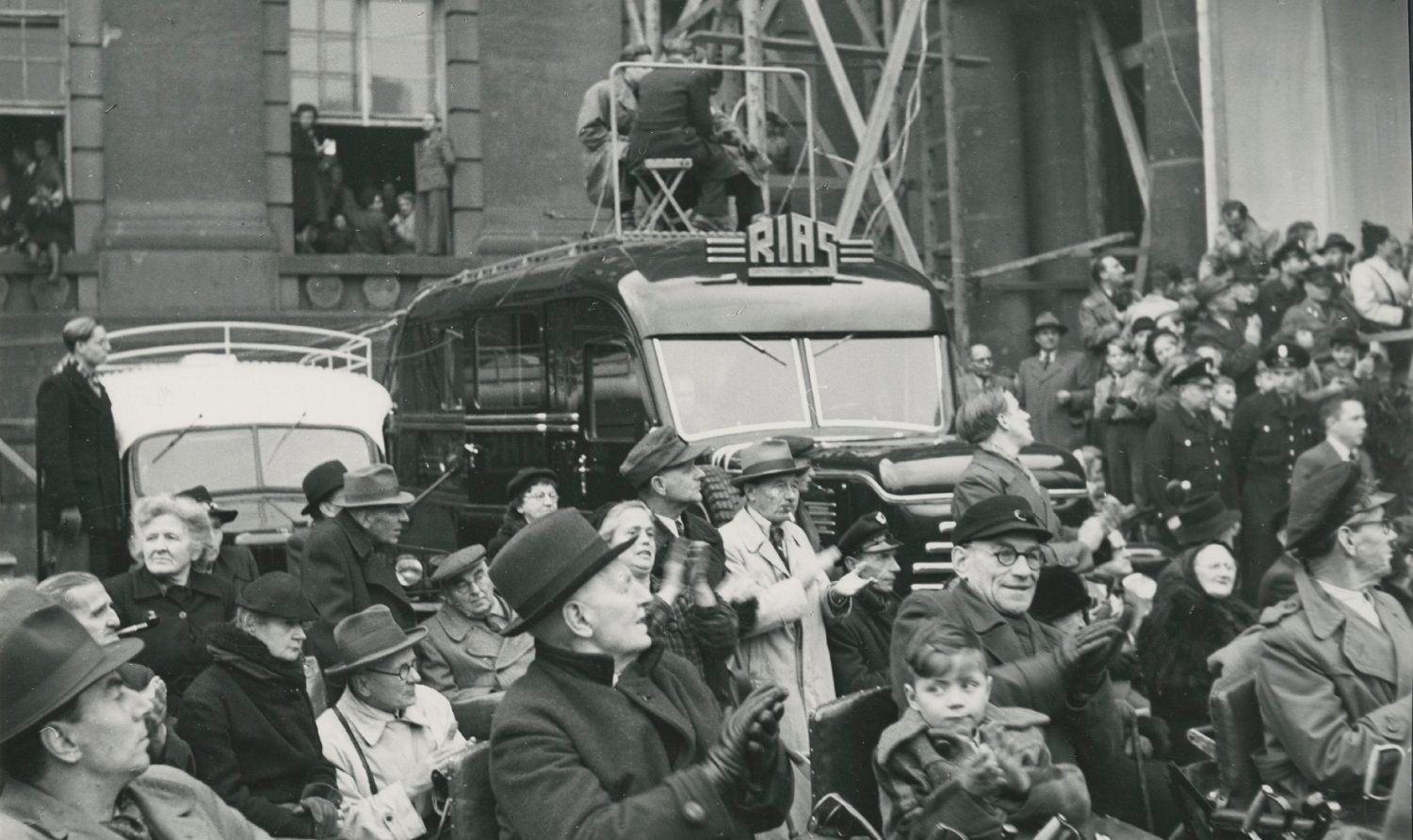 Enlarge photo: A crowd applauds, in the background there is a car with the inscription RIAS. 