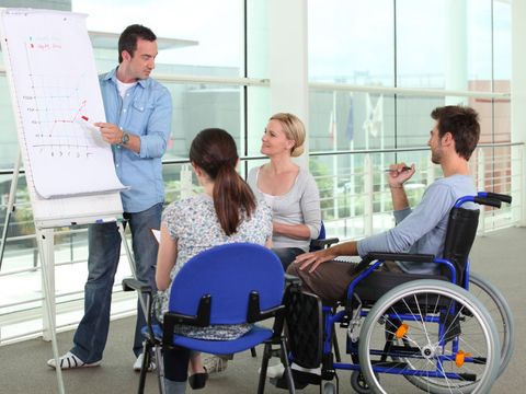 Besprechung unter 4 MItarbeitern, zwei sitzen auf einem Stuhl, einer im Rollstuhl und der vierte steht an einem Flipchart und erklärt ein Diagramm 