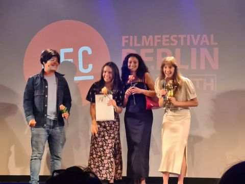 Bildvergrößerung: Vier Jugendliche stehen zusammen auf einer Bühne. Sie halten Rosen, eine Auszeichnung und einen goldenen Preis in der Hand.