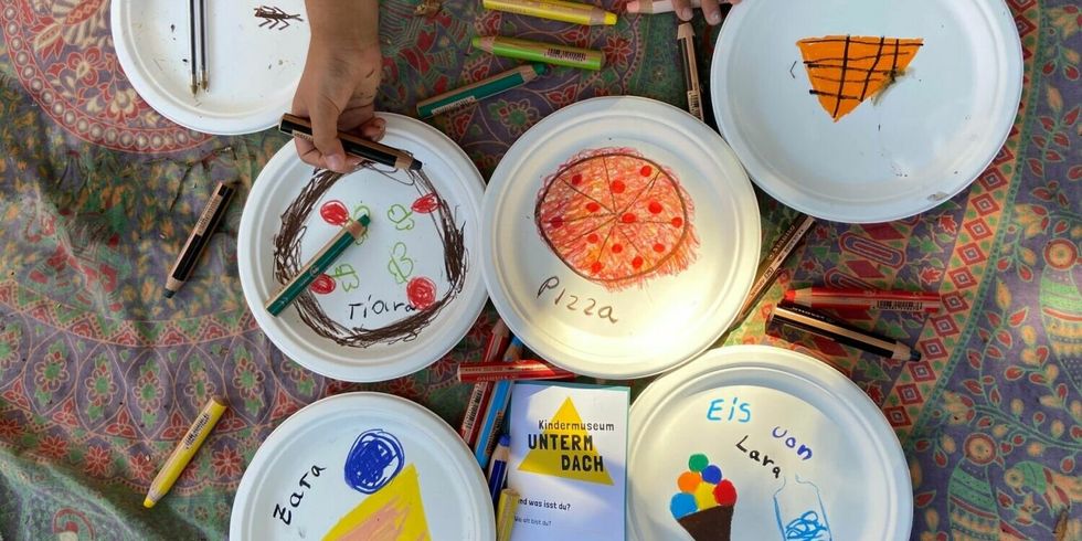 Von Kindern mit Motiven von Essen aus der ganzen Welt gestaltete Pappteller auf bunter Tischdecke.