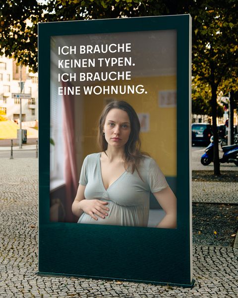 Auf einer Stadtwerbetafel ist eine schwangere Frau abgebildet mit dem Text: “ICH BRAUCHE KEINEN TYPEN. ICH BRAUCHE EINE WOHNUNG.” Im Hintergrund sind Bäume, Gebäude und ein Gehweg zu sehen.