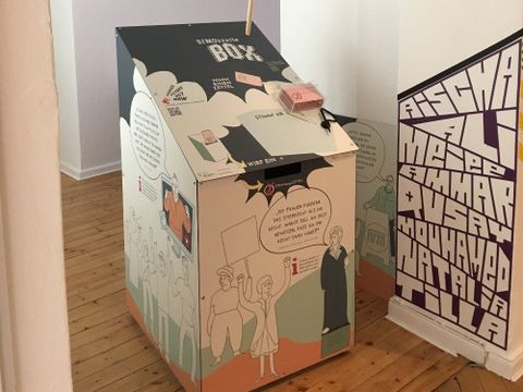 Bildvergrößerung: Eine bunte Box steht mit einem Schild mit der Aufschrift "Demokratie-Box" in einem Raum. Auf der Box ist ein Behältnis mit Zetteln und an der Seite ist ein Schlitz.