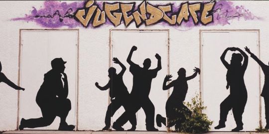 Schwarze Figuren, die tanzen mit Graffiti an der Wand im Hinergrund mit dem Text: Jugendcafe