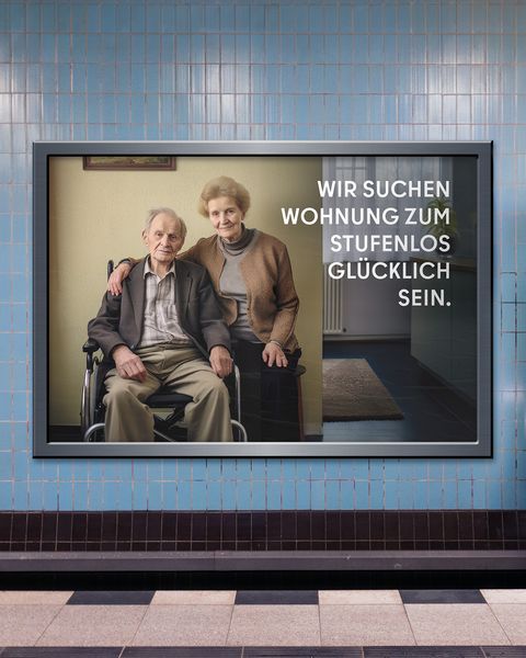 Auf dem Bild ist eine Werbetafel in einer U-Bahn-Station zu sehen. Die Tafel zeigt ein älteres Paar, wobei der Mann im Rollstuhl sitzt. Der Text auf der Tafel lautet: “WIR SUCHEN WOHNUNG ZUM STUFENLOS GLÜCKLICH SEIN.” Im Hintergrund bewegt sich ein Zug.