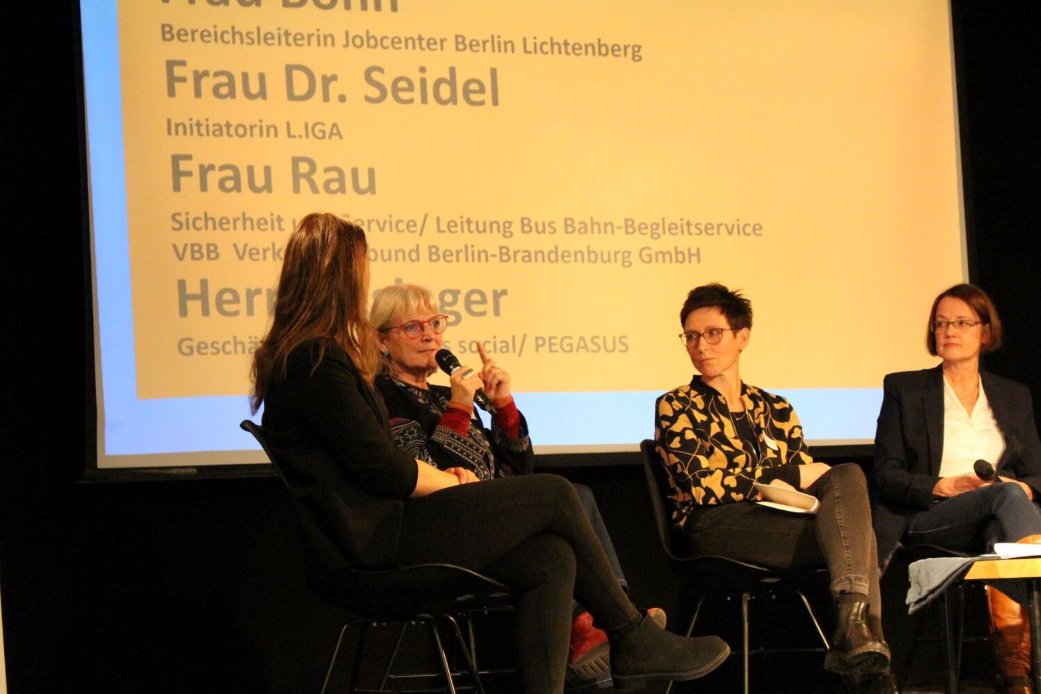 Vier Frauen sitzen in einem Diskussionspodium. Eine farbige Präsentation im Hintergrund zeigt einen Ausschnitt auf Namen: Frau Dr. Seidel, Frau Rau.