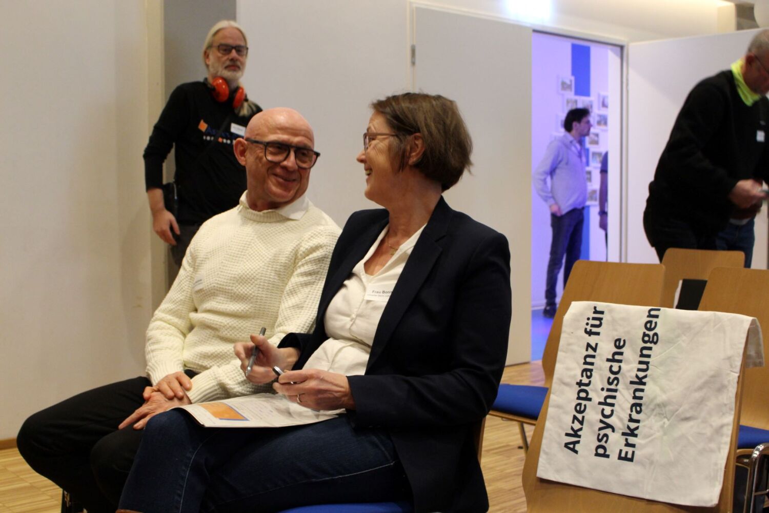 Ein Mann und eine Frau sitzen in einer Reihe von Stühlen und unterhalten sich fröhlich. Auf einem leerem Stuhl neben ihnen liegt ein Stoffband mit der Aufschrift: Akzeptanz für psychische Erkrankungen.