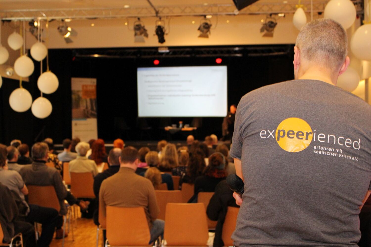 In einem Saal sitzen viele Menschen vor einer Bühne. Im Hintergrund ist ein Bildschirm zu sehen. Ein Mann blickt rückwärtsgewandt auf die Menschen. Sein T-Shirt trägt die Aufschrift: expeerienced.