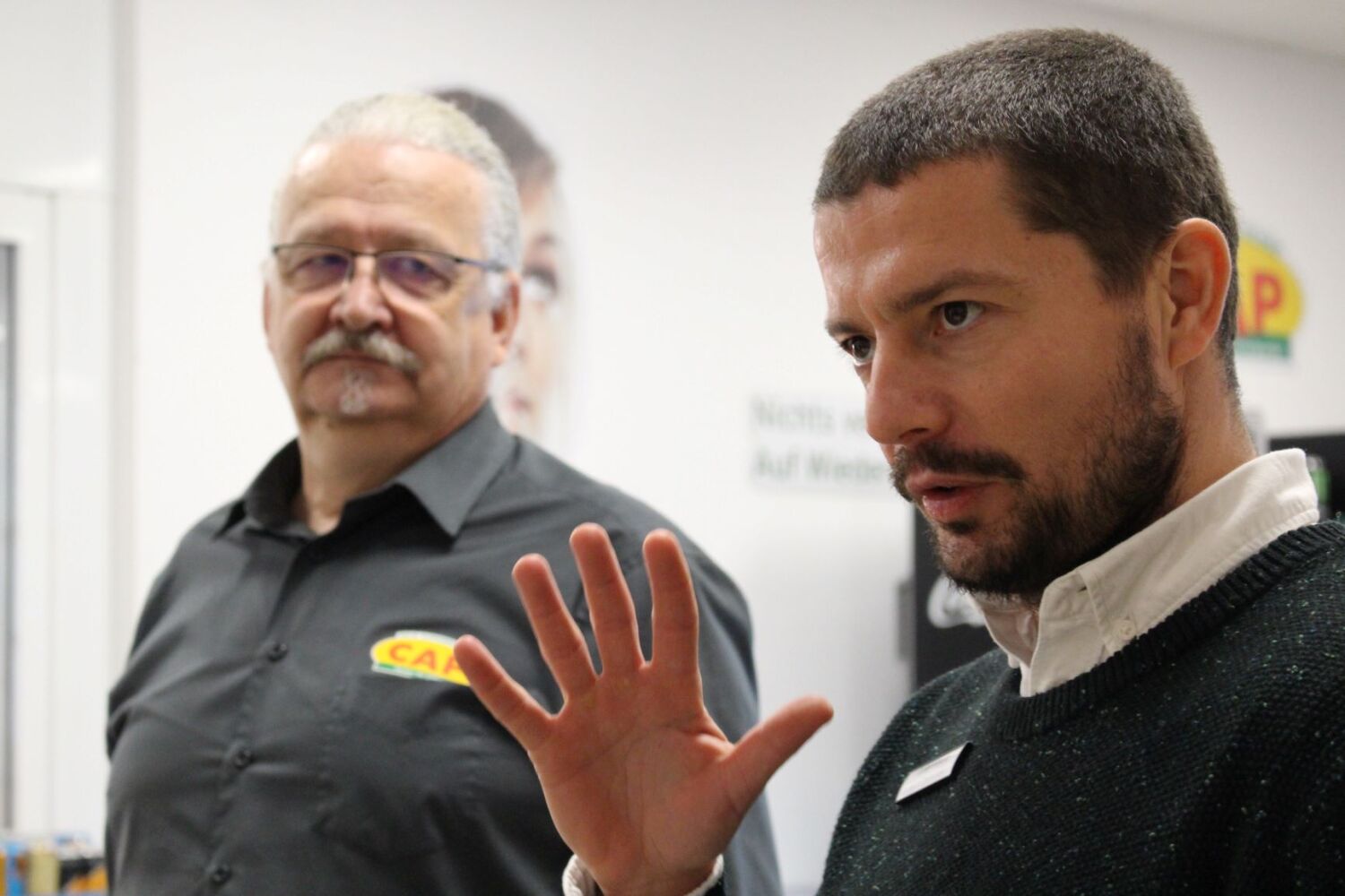 Ein Mann spricht und gestikuliert mit seiner Hand. Ein Mann im Hintergrund hört ihm aufmerksam zu.