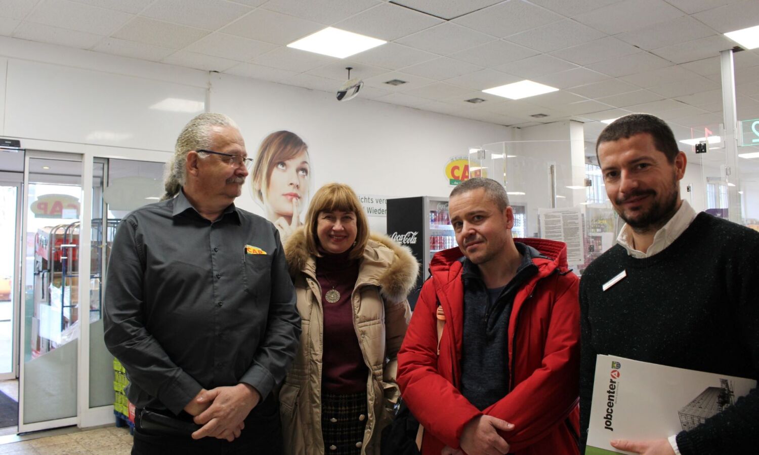 Eine Gruppe von vier Menschen stehen zusammen in einem Supermarkt und schauen freundlich in die Kamera.