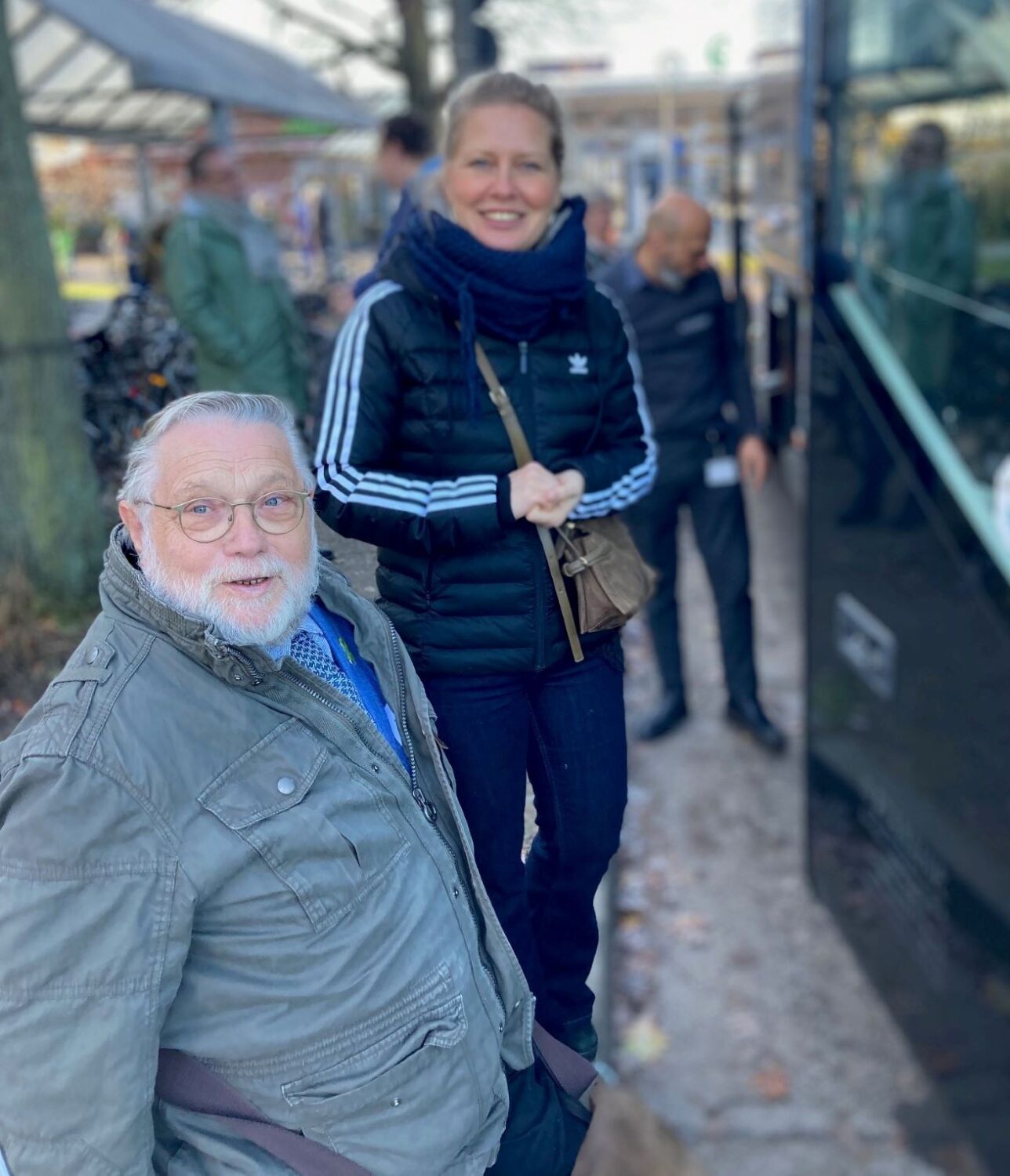 Eine Frau und ein Mann schauen in die Kamera. Ein schwarzer Bus ist im Hintergrund erkennbar.