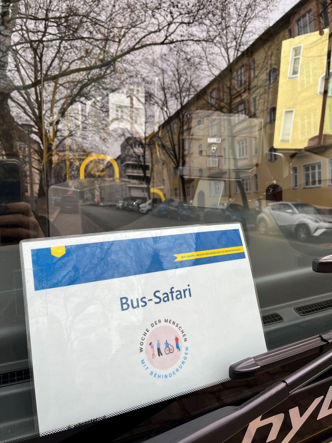 Ein Schild mit der Aufschrift Bus-Safari und einem buntem Logo Woche der Menschen mit Behinderungen klemmt hinter einer Busscheibe.