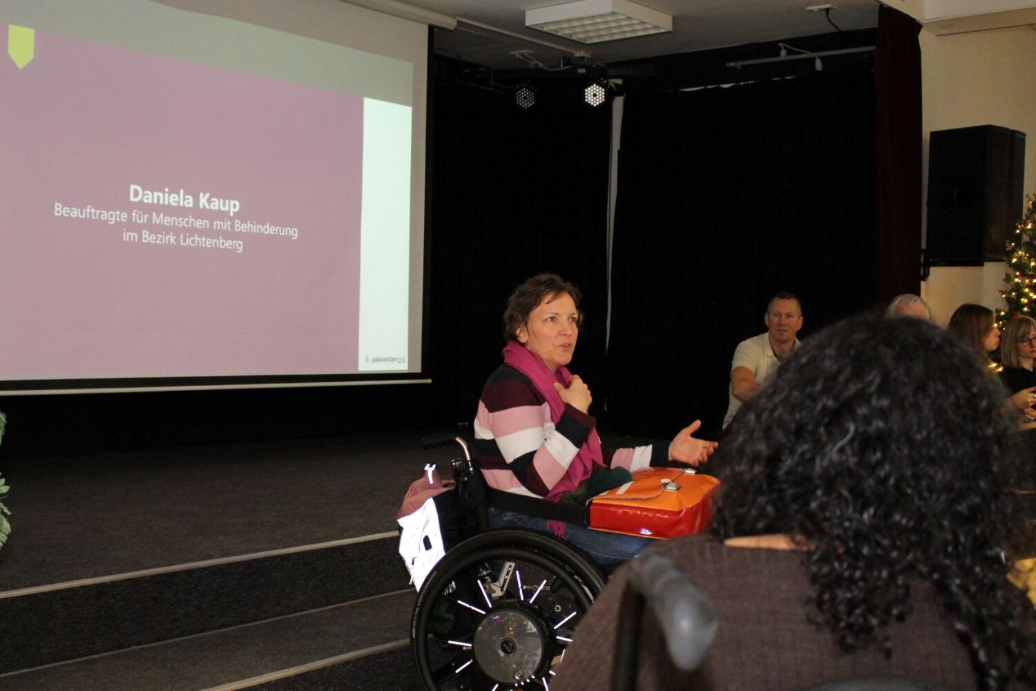 Eine Frau im Rollstuhl spricht zu einer Gruppe von sitzenden Menschen. Eine farbige Präsentation im Hintergrund zeigt ihren Namen: Daniela Kaup.