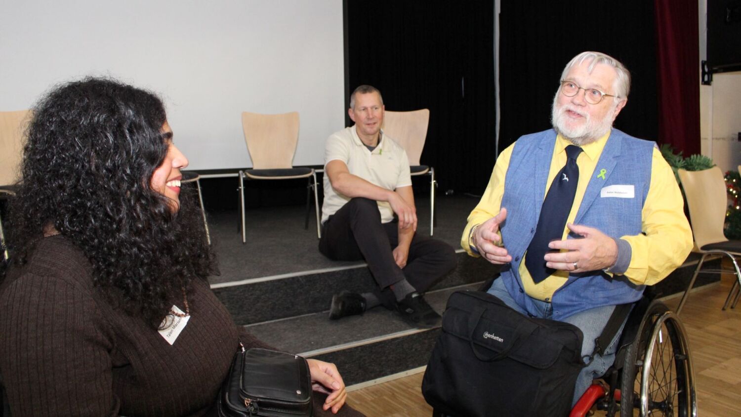 Eine Frau und ein Mann im Rollstuhl unterhalten sich. Ein dritter Mann sitzt auf einer niedrigen Bühne hinter den beiden und hört ihnen zuz.